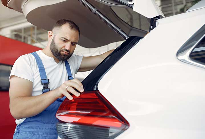 A man in blue overalls working on the back of a white car.